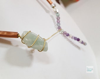 May include: A necklace featuring a polished light green stone wrapped in gold-colored wire, connected to a copper tube and a strand of small purple and green beads. The bottom right corner displays the text Temp LAB in a small white square.