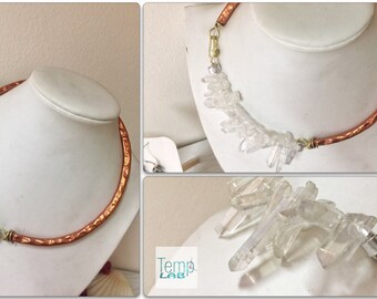 May include: A necklace featuring a hammered copper collar connected to a cluster of clear, raw quartz crystals. The piece includes brass wire wrapping and a metal clasp. A small square logo with the text Temp LAB is visible in the corner.