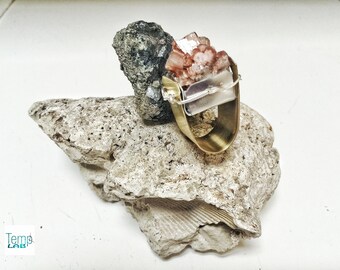 May include: A ring featuring a wide, polished brass band and a raw, pinkish-brown crystal cluster secured with silver-toned wire. The ring is positioned against a dark, textured mineral and a larger, porous, light-colored rock base.