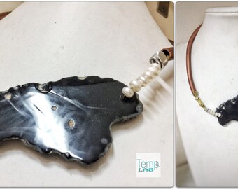 May include: A necklace featuring a large, irregular, polished black stone pendant with white inclusions, suspended from a copper-toned cord. The design includes white pearl accents, gold-toned wire wrapping, and a silver-toned metal nut detail.