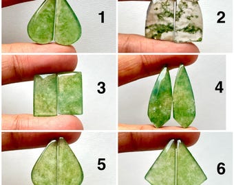 May include: Six pairs of polished gemstones in various shapes and shades of green. The gemstones are held between fingers, showcasing their different cuts: heart, rectangular, teardrop, and geometric shapes. The stones have a translucent quality.