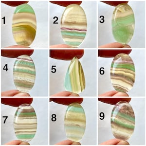 Può includere: Nove pietre di fluorite lucidata, presentate in varie forme e colori. Le pietre sono ovali e a goccia, con bande di tonalità verdi, gialle, viola e chiare. Ogni pietra è tenuta tra le dita, mostrando le variazioni naturali e la traslucenza del minerale.