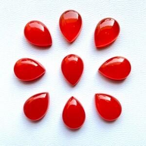 May include: Eight vibrant red teardrop-shaped cabochons. These polished gemstones have a glossy finish and are arranged on a white surface. The cabochons are likely used for jewelry making or other craft projects.