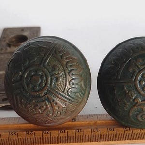 May include: Two round, dark bronze door knobs featuring intricate embossed floral and geometric patterns, accompanied by two matching rectangular backplates. A wooden ruler is positioned in front, showing the knobs are approximately 2.25 inches wide.