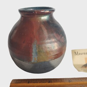 May include: A small, handcrafted ceramic vase with a rounded body and a narrow opening. The vase features a blend of colors, including red, brown, and blue, with a metallic sheen. The base is a darker gray. A wooden ruler is at the bottom.
