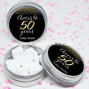 Könnte beinhalten: Drei kleine, silberfarbene Dosen mit Deckeln. Jede Dose hat ein schwarzes Etikett mit goldenem Text, der "Cheers to 50 years" und den Namen "Libby Smith" zeigt. Eine Dose ist geöffnet und zeigt herzförmige Süßigkeiten.