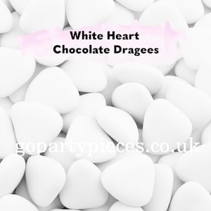 Op de afbeelding: Close-up van witte hartvormige chocoladedragees. De snoepjes zijn verspreid, waardoor een textuurpatroon ontstaat. De tekst "White Heart Chocolate Dragees" wordt in het midden bovenaan weergegeven.