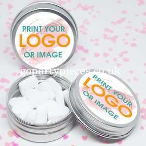 Puede incluir: Dos latas de plata con tapas. Las tapas tienen etiquetas rosas y blancas con purpurina dorada. Las etiquetas dicen "Imprime tu logotipo o imagen". Una lata está abierta y contiene mentas blancas.