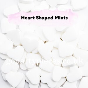 Op de afbeelding: Een close-up van witte hartvormige pepermuntjes verspreid over een witte ondergrond. De afbeelding bevat de tekst "Heart Shaped Mints". Perfect voor feesten en evenementen.