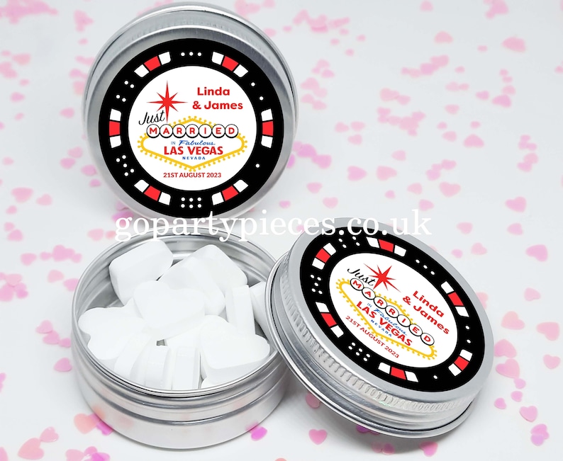 Op de afbeelding: Drie kleine, zilveren blikken met deksels met een zwart en rood ontwerp en de tekst "Just Married Fabulous Las Vegas Nevada" met een datum. E&eacute;n blik is open en toont hartvormige snoepjes.