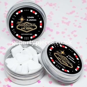 May include: Two silver tins with black lids. The lids have a red and white border and a black and gold design that says "Just Married in Fabulous Las Vegas" with the date "21st August 2023". The text "Linda & James" is above the Las Vegas design. One tin is open and contains white mints.