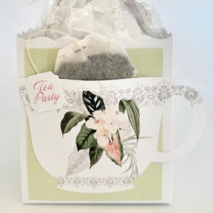 May include: A tea party favor with a tea bag, packaged in a white and green paper cup-shaped container. The container has a floral design and the words "Tea Party". A tag reads "Thank You for showering the beau-tea-ful Bride to Be with love".