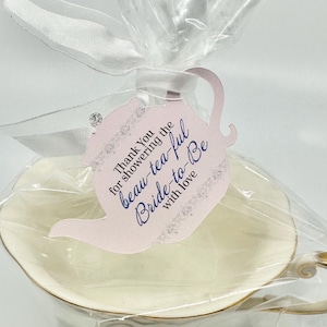 May include: A cream-colored teacup with a gold rim and handle, wrapped in clear cellophane with a white ribbon. A light pink teapot-shaped tag is attached, featuring the text: Thank You showering the beau-tea-ful Bride-to-Be with love.