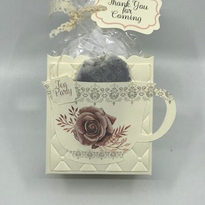 May include: A white teacup-shaped box with a brown rose and leaves design. The box contains a tea bag and a small white gift bag with a tag that says "Thank You for Coming". The box also has a tag that says "Tea Party".