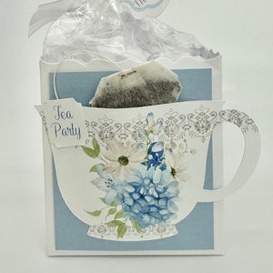 May include: A blue and white teacup favor box with a tea bag inside. The box has a scalloped edge and a floral design. The tag attached to the box says "Thank You for showering the beau-tea-ful Mother-to-Be with love".