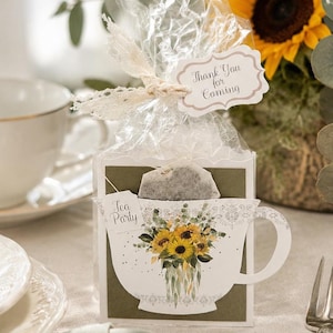 Puede incluir: Una caja de regalo cuadrada que contiene una bolsita de té, envuelta en plástico transparente con una cinta de encaje y una etiqueta que dice Thank You for Coming. La caja muestra un gráfico de una taza con girasoles y el texto Tea Party.