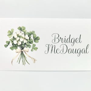 May include: A white card with a watercolor illustration of a bouquet of white flowers and green shamrocks tied with a beige ribbon. The card has the name "Bridget McDougal" in elegant script.