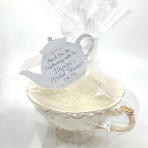 May include: A wrapped teacup and saucer set, likely a bridal shower favor. The teacup is cream-colored with gold accents and a delicate floral design. A decorative tag in the shape of a teapot reads "Thank You for Celebrating with Us Marisa's Bridal Shower 3.26.2026."