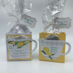 May include: Two yellow teacup-shaped boxes with a tea bag inside, decorated with lemons and flowers. The boxes have a tag that says "Thank You for Coming" and "Tea Party". The boxes are for a bridal shower.