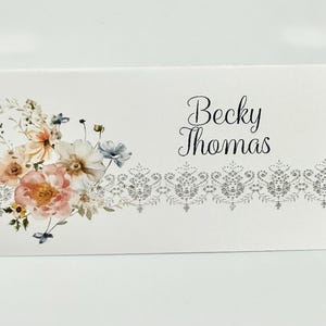 May include: A white card with floral watercolor art and the name "Becky Thomas" in elegant script. The floral arrangement features peach, pink, and blue flowers. A decorative silver border accents the bottom.