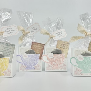 May include: Gift sets featuring tea bags and decorative teacup-shaped boxes. Each box has a different pastel color teacup design and a small tag with a quote. The sets are wrapped in clear cellophane and tied with lace ribbon.