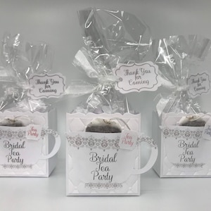 May include: Three white boxes with a teacup design, each containing a tea bag and a tag that says "Thank You for Coming". The boxes are decorated with a silver lace pattern and the words "Bridal Tea Party".