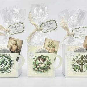 May include: Three white favor boxes shaped like teacups with green and gold designs. Each box has a tea bag, a small card with a leprechaun or a woman in a green dress, and a tag that says "Thank You for Coming".