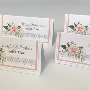 May include: Four white place cards with pink roses and a silver lace design. The cards have the names Karen Boisseau, Amy Grigoriou, Lorelei Sutherford, and Adele Frost, each with the text "Table One".
