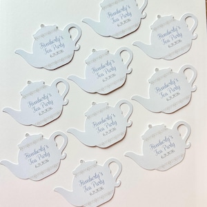 May include: White die-cut teapot-shaped party favors. Each teapot has the text "Kimberly's Tea Party 6.21.2026" in blue script. The teapots are decorated with a delicate floral pattern and a small gem on the lid.