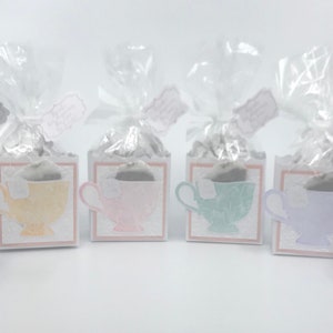 May include: Six small white boxes with teacup designs in various pastel colors, each box is wrapped in clear cellophane and tied with a ribbon. The boxes are arranged in a row on a white surface.
