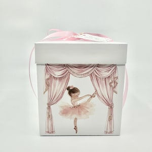 May include: A white square gift box with a pink ribbon and tag. The box features a watercolor illustration of a ballerina on stage, wearing a pink tutu and pointe shoes, with pink curtains in the background.