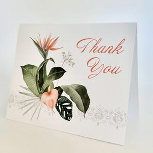 May include: A folded white card with a watercolor floral design and the words "Thank You" in a copper-colored script. The floral arrangement includes a bird of paradise flower, green leaves, and a peach-colored anthurium.