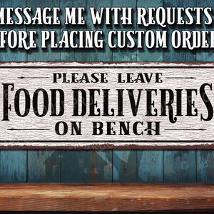 May include: White metal sign with black text that reads "Please Leave Food Deliveries On Bench". The sign has a distressed, vintage look and is mounted on a blue wooden wall.