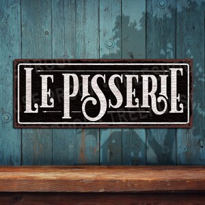 May include: A black metal sign with white lettering that reads "LE PISSERIE". The sign has a distressed, vintage look and is hanging on a blue wooden wall.