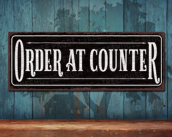 May include: A rectangular black sign with white text that reads "ORDER AT COUNTER." The sign has a distressed, rustic look with a brown border. The background is a weathered blue wooden wall.