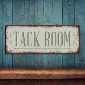 May include: A rectangular metal sign with the words "TACK ROOM" in gray lettering. The sign has a distressed, vintage look with a white background and a brown border. The sign is mounted on a weathered blue wooden wall.