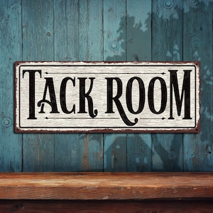 May include: A white metal sign with black lettering that reads "TACK ROOM". The sign has a rustic, distressed look and is hanging on a blue wooden wall.