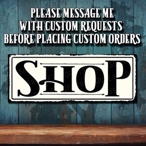 May include: A white sign with black text that reads "PLEASE MESSAGE ME WITH CUSTOM REQUESTS BEFORE PLACING CUSTOM ORDERS" and "SHOP". The sign is on a blue wooden background.