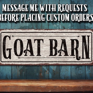May include: A rustic white sign with black lettering that reads "GOAT BARN". The sign is hanging on a blue wooden wall. The sign is made of metal and has a distressed look.