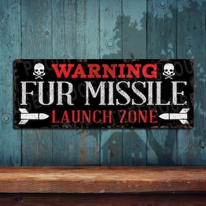 May include: A black and white sign with red text that reads "WARNING FUR MISSILE LAUNCH ZONE". The sign has two skull and crossbones symbols on either side of the text. The sign is made of metal and has a distressed look.