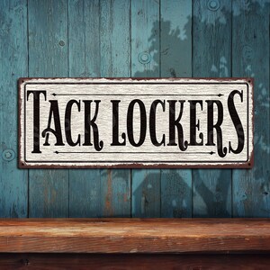 May include: A white metal sign with black lettering that reads "TACK LOCKERS" against a blue wooden background.