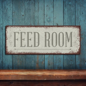 May include: A white metal sign with the words "FEED ROOM" in gray lettering on a blue wooden background. The sign has a distressed look and is hanging on a wooden wall.