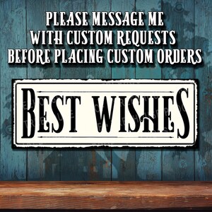 May include: A rectangular sign with the words "BEST WISHES" in bold black letters. The sign has a white background with a black border and is set against a blue wooden background. The text "PLEASE MESSAGE ME WITH CUSTOM REQUESTS BEFORE PLACING CUSTOM ORDERS" is above the sign.