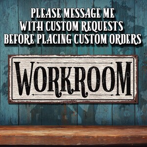 May include: A white sign with black text that reads "WORKROOM" on a rustic blue wood background. The sign has a distressed look and is attached to the wall with screws. The text above the sign reads "PLEASE MESSAGE ME WITH CUSTOM REQUESTS BEFORE PLACING CUSTOM ORDERS."