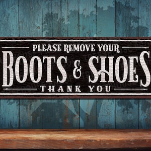 May include: A rustic black and white sign with the text "Please remove your boots & shoes thank you" on a wooden background.