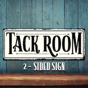 May include: A white metal sign with black lettering that reads "Tack Room" with a distressed look. The sign is on a blue wooden background and has the text "2 - Sided Sign" below it.