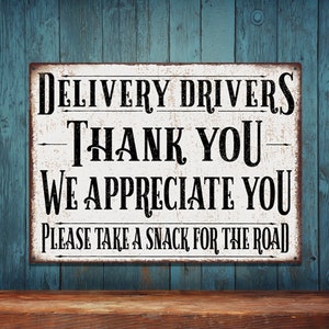 May include: A white metal sign with black text that reads "DELIVERY DRIVERS THANK YOU WE APPRECIATE YOU PLEASE TAKE A SNACK FOR THE ROAD". The sign is hanging on a blue wooden wall.