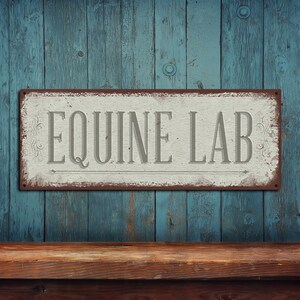 May include: A rectangular metal sign with the words "EQUINE LAB" in gray letters. The sign has a distressed, vintage look with a white background and a brown border. The sign is mounted on a blue wooden wall.
