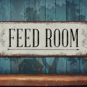May include: A white sign with brown edges and distressed paint reads "FEED ROOM" in black lettering. The sign has decorative flourishes on either side of the text and a small arrow pointing to the right. The sign is hanging on a blue wooden wall.