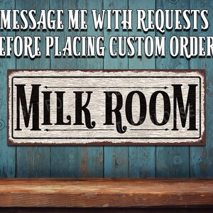 May include: A white sign with black text that reads "Milk Room" on a blue wooden background. The sign has a distressed, vintage look. The text is in a decorative font with arrows pointing to the left and right.